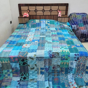 May include: A patchwork quilt featuring a variety of blue, teal, and turquoise fabric squares with diverse floral, geometric, and paisley patterns. The quilt is stitched with visible running thread lines and covers a bed with a wooden headboard.