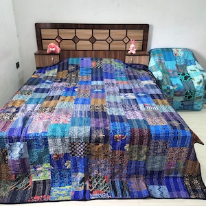 May include: A large rectangular quilt featuring a patchwork design of various blue, teal, and navy fabric squares with intricate patterns and visible stitching, covering a bed and a matching chair in a bedroom setting.