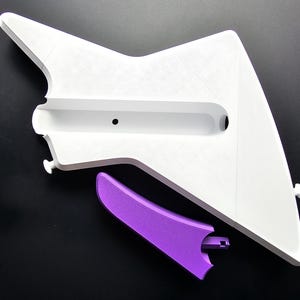 Puede incluir: Un objeto blanco angular con una ranura rectangular larga y un pequeño agujero redondo. Una pieza curva púrpura se coloca debajo. Los objetos son probablemente componentes de un artículo más grande, posiblemente una pieza decorativa o funcional.