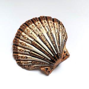 May include: A white seashell painted with black and gold geometric patterns. The shell is closed and has a smooth, glossy finish.