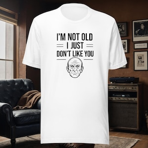 Op de afbeelding: Wit T-shirt met zwarte tekst die "Ik ben niet oud = Ik ben gewoon = Vind je niet leuk" luidt. Onder de tekst staat een cartoonillustratie van een ouder man.