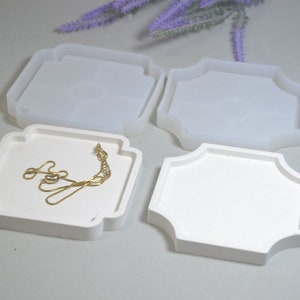 May include: Four white silicone moulds for resin casting. The moulds are shaped like trays with a decorative edge. One mould has a gold chain inside.