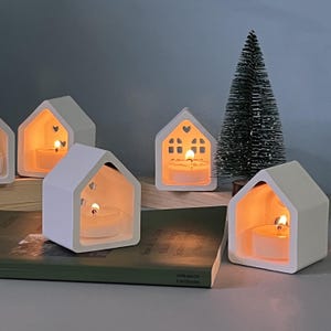 May include: White ceramic house-shaped candle holders with lit tea lights inside. The candle holders have cut-out windows and a heart-shaped opening. A small, green Christmas tree is in the background. The candle holders are on a table.