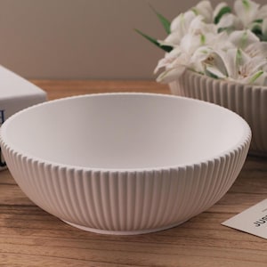 Op de afbeelding: Een witte keramische kom met een geribbeld ontwerp. De kom is leeg en staat op een houten oppervlak.