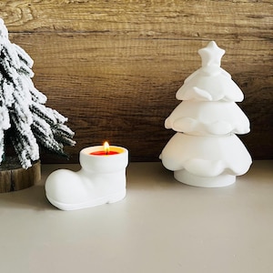 May include: White ceramic candle holders shaped like a Christmas tree and a Santa boot. The boot has a red candle inside.