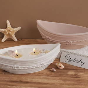 Op de afbeelding: Twee witte keramische kaarshouders in de vorm van boten met touwdetails. De boten zijn gevuld met witte was en hebben twee brandende kaarsen. De boten staan op een lichtbruin oppervlak met een kleine schelp en een bord met de tekst "Yesterday".