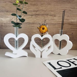 Puede incluir: Tres jarrones blancos en forma de corazón con tubos de vidrio, cada uno con diferentes elementos decorativos. Uno tiene un recorte en forma de corazón y una base en forma de mano. Otro tiene un recorte en forma de corazón con un diseño en forma de mano.