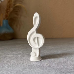 Puede incluir: Figurita de clave de sol de cerámica blanca. La nota está de pie sobre una pequeña base.