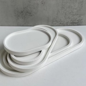 Könnte beinhalten: Ein Satz aus vier weißen, ovalen Keramiktabletts, die übereinander gestapelt sind. Die Tabletts haben eine glatte, matte Oberfläche.