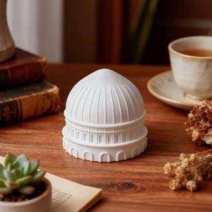 May include: A white, dome-shaped decorative item with architectural detailing, placed on a wooden surface. The design features vertical lines and arched features. The background includes a teacup, books, and a small succulent plant.