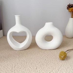 Puede incluir: Dos jarrones de cerámica blanca con diseños únicos de recortes. Un jarrón tiene una abertura en forma de corazón, mientras que el otro tiene una abertura circular.