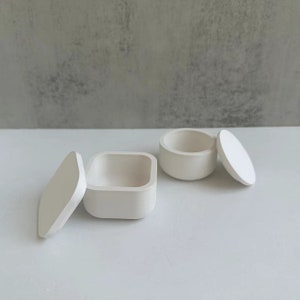 Peut inclure: Deux petits contenants en céramique blanc cassé avec couvercles. L'un est carré, l'autre est rond. Les couvercles sont légèrement entrouverts, révélant l'intérieur des contenants. Les contenants sont sur une surface blanche.