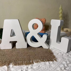 Könnte beinhalten: Weiße Holzbuchstaben, die "A & L" auf einer Jutestoff-Oberfläche buchstabieren. Die Buchstaben sind freistehend und haben ein schlichtes, modernes Design.