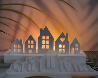 May include: A white, decorative candle holder shaped like a row of houses. Each house has cut-out windows, and one has a heart-shaped cutout. The candle holder sits on a base with a mountain-like design, with a warm glow emanating from within.