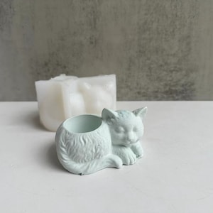 Può includere: Un vaso da fiori a forma di gatto addormentato di colore azzurro chiaro con un'apertura rotonda per la semina. Il vaso è in cemento e ha una superficie testurizzata.