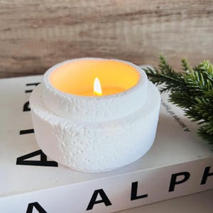 Puede incluir: Portavelas blanco con superficie texturizada y una vela parpadeante en su interior. El portavelas está sobre un libro blanco con la palabra "ALPHA" impresa en la portada.