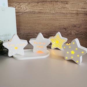 May include: Four white star-shaped decorative lights with yellow internal illumination. Two stars have text, one reads "Für Dich" and the other "Familien Fest". The lights sit on a white rectangular base.