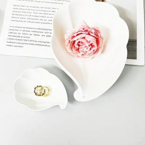 Puede incluir: Dos platos de cerámica blanca con forma de hoja. El plato más grande tiene una flor rosa en la parte superior. El plato más pequeño contiene dos anillos dorados.