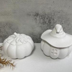 Puede incluir: Dos recipientes de cerámica blanca con forma de calabaza. Un recipiente tiene una tapa con forma de sombrero de bruja y una pequeña ventana. El otro recipiente tiene un diseño de hoja en la tapa.