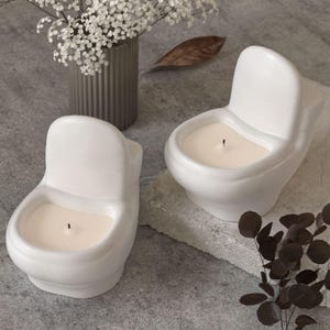 May include: Two white ceramic toilet-shaped candles with a creamy wax fill and a single wick. The candles are placed on a grey surface, with a vase of white flowers and decorative foliage in the background. The candles are a unique home decor item.