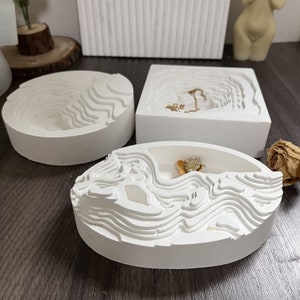 May include: Three white ceramic trinket dishes with a wavy, textured design. The dishes are different shapes and sizes, and one has a gold ring inside.