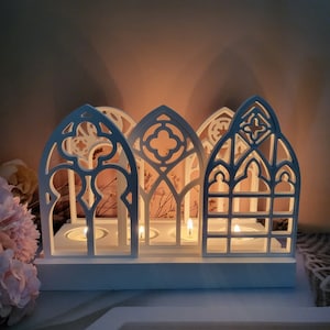 May include: White candle holder with a gothic arch design. The holder has three candle slots and is made of wood. The candle holder is lit with three candles.