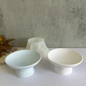 Puede incluir: Tres cuencos decorativos pequeños con bases de pedestal. Los cuencos están hechos de un material blanco y tienen una forma redonda y lisa. Los cuencos están dispuestos en una fila sobre un fondo gris.