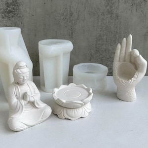 May include: A collection of white resin molds and finished products. The items include a seated Buddha statue, a lotus flower base, a hand-shaped candle holder, and various cylindrical molds. The objects are arranged on a white surface.