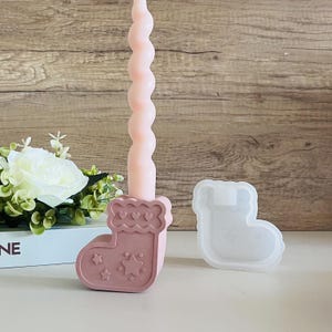 Puede incluir: Portavelas de cerámica rosa con forma de calcetín de Navidad con una vela blanca en espiral. El portavelas tiene un diseño de corazones y estrellas. También se muestra un molde de silicona del calcetín.