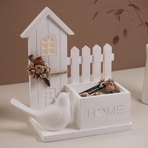 Może przedstawiać: Biała, dekoracyjna ozdoba do domu z figurką ptaka, miniaturowym domkiem z oknem i małym płotkiem. Słowo "HOME" jest wyryte na froncie małego pudełka. Suszone kwiaty i wazon dopełniają ekspozycję.