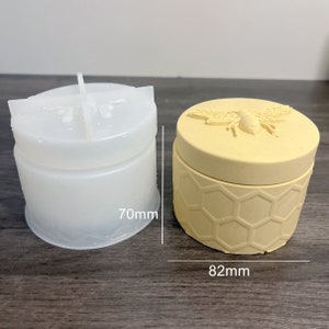 Puede incluir: Un molde de silicona blanco y un tarro de cerámica beige con un diseño de abeja en la tapa. El tarro tiene un patrón de panal en el lateral. El molde tiene un diámetro de 70 mm y el tarro tiene un diámetro de 82 mm.