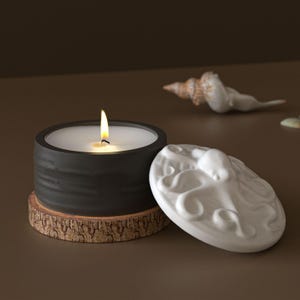May include: A lit black candle in a round container with a white lid featuring an octopus design. The candle sits on a small wooden coaster. A seashell is in the background.