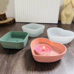 Puede incluir: Cuatro portavelas de silicona en diferentes formas y colores. Un cuadrado verde, un cuadrado blanco, un corazón blanco y un corazón rosa. El portavelas en forma de corazón rosa tiene una pequeña vela blanca dentro.