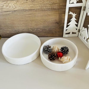 May include: Two white ceramic bowls with a ribbed design. The smaller bowl is filled with pine cones and small white flowers. A red ornament is also in the bowl.