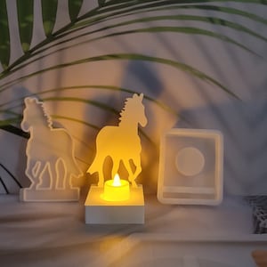 Puede incluir: Tres moldes de silicona blancos para hacer velas. Los moldes tienen forma de caballos y un cuadrado con una hendidura redonda. Una vela encendida está en uno de los moldes de caballo.
