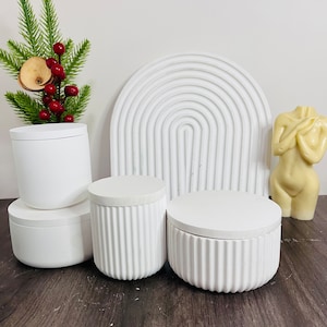 May include: Four white cylindrical containers with lids, two of which have a ribbed texture. The containers are arranged on a wooden surface in front of a white, textured, arched background.
