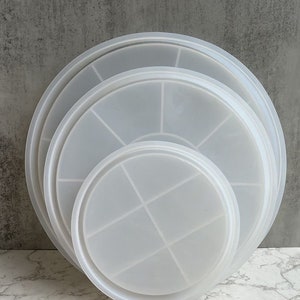 May include: Three clear plastic round cake pans with a grid pattern on the bottom. The pans are stacked on top of each other, with the largest pan on the bottom and the smallest pan on top.