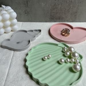 Puede incluir: Tres bandejas decorativas con forma de concha, corazón y almeja. La bandeja con forma de almeja tiene un esmalte verde y está llena de cuentas de perlas blancas. La bandeja con forma de concha es gris y la bandeja con forma de corazón es rosa.
