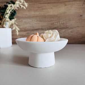 May include: White ceramic pedestal dish with a wide, shallow bowl and a round base. The dish is decorated with a small orange pumpkin and a white flower.