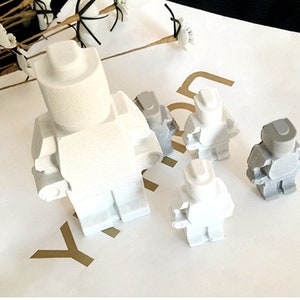 以下が含まれることがあります： 白とグレーの小さなロボットの置物5つ。一番大きなロボットは白で丸い頭をしている。他の4つのロボットは小さく、頭の色がそれぞれ違う。