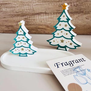 May include: Two white paper Christmas trees with gold stars and green outlines on a white oval tray. A card with the word "Fragrant" and a small brown circle is on the tray.