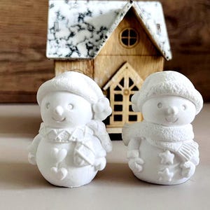 Könnte beinhalten: Zwei weiße Schneemann-Figuren aus Keramik mit bemalten Details. Ein Schneemann trägt einen Schal mit Herzen und der andere einen Schal mit Sternen.