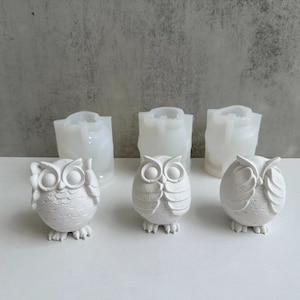 Pode incluir: Três estatuetas de coruja brancas, cada uma com uma pose diferente, sentadas em uma superfície cinza. As corujas são feitas de um material branco liso e têm detalhes precisos, incluindo olhos, bicos e penas. Atrás das corujas, há três moldes de silicone transparentes.