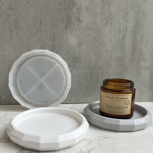 May include: Two white ceramic candle holders, one with a geometric design and the other with a simple round shape. A brown glass candle jar with a label that reads "Lavender Eucalyptus Soy Wax Candle Hand Poured" is sitting on the geometric candle holder.