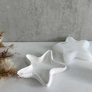 May include: Two white ceramic starfish-shaped dishes. The dishes are identical in shape and size, but one is slightly raised and appears to be a lid for the other.