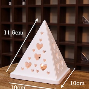 Può includere: Portacandele in ceramica bianca a forma di piramide con tagli a forma di cuore. Il portacandele misura 11,5 cm di altezza, 10 cm di larghezza e 10 cm di profondità.