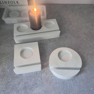 Könnte beinhalten: Vier graue Beton-Kerzenhalter. Zwei Halter sind rechteckig mit einer einzelnen Kerzenvertiefung, einer ist rechteckig mit zwei Kerzenvertiefungen und einer ist rund mit einer einzelnen Kerzenvertiefung.
