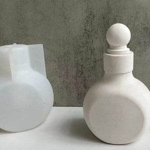 Könnte beinhalten: Eine weiße Keramikflasche mit rundem Körper und rundem Deckel. Die Flasche hat eine glatte, matte Oberfläche.