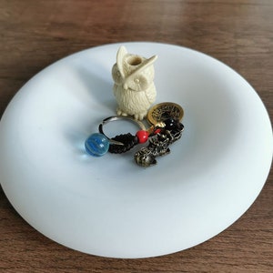 Puede incluir: Un plato de cerámica blanco con una pequeña figurita de búho blanco, una cuenta de vidrio azul, una moneda de oro y un amuleto negro y dorado.