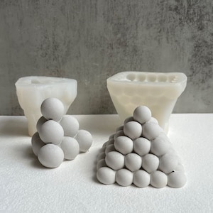 Könnte beinhalten: Zwei weiße Betonformen zum Herstellen von dekorativen Kugeln. Eine Form ist eine einzelne, große, schalenförmige Form. Die andere Form ist eine pyramidenförmige Form mit mehreren kleinen Kugelvertiefungen.